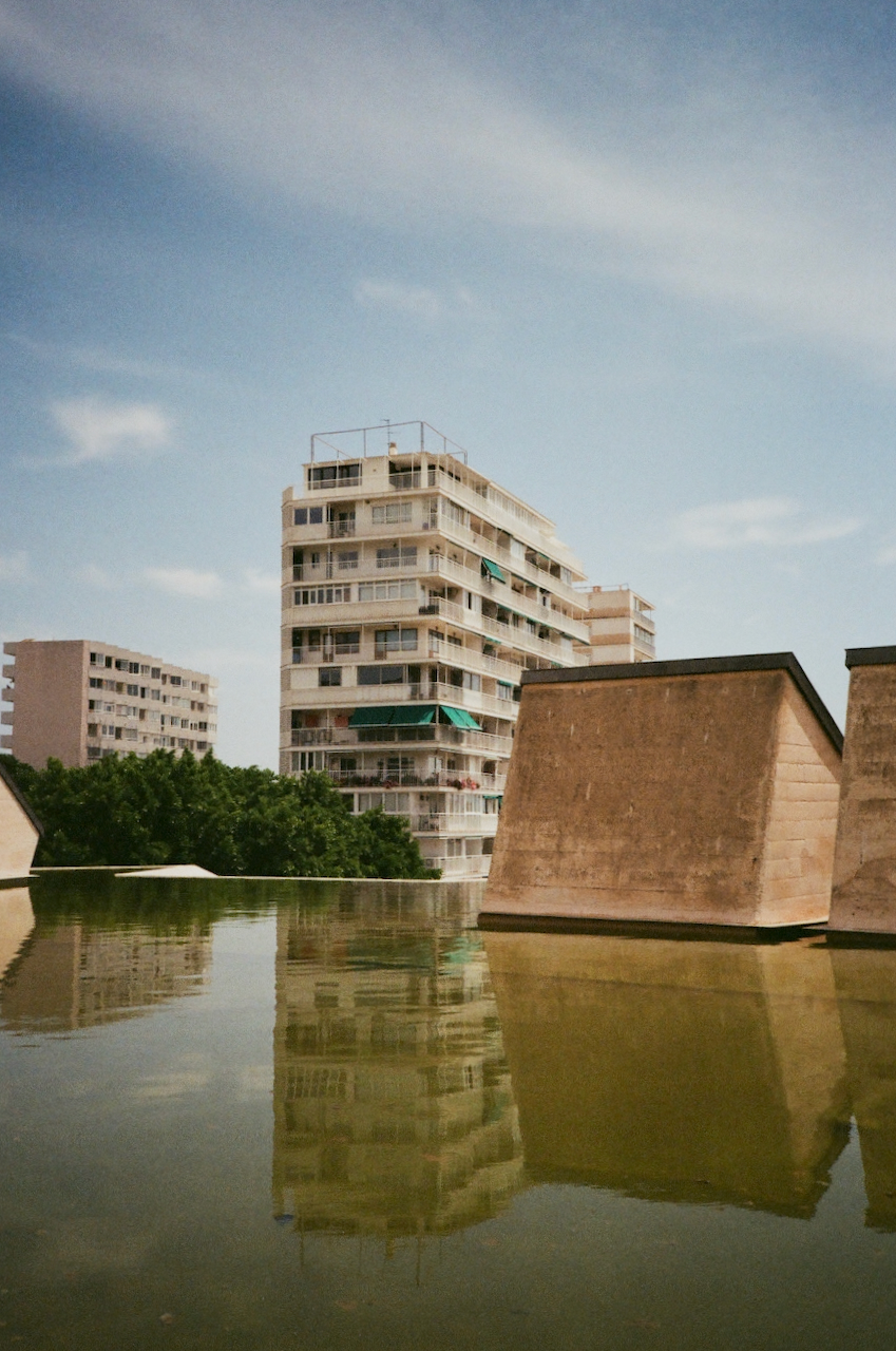 A serene urban scene featuring a reflection of a building in a body of water, with a clear blue sky and a few clouds