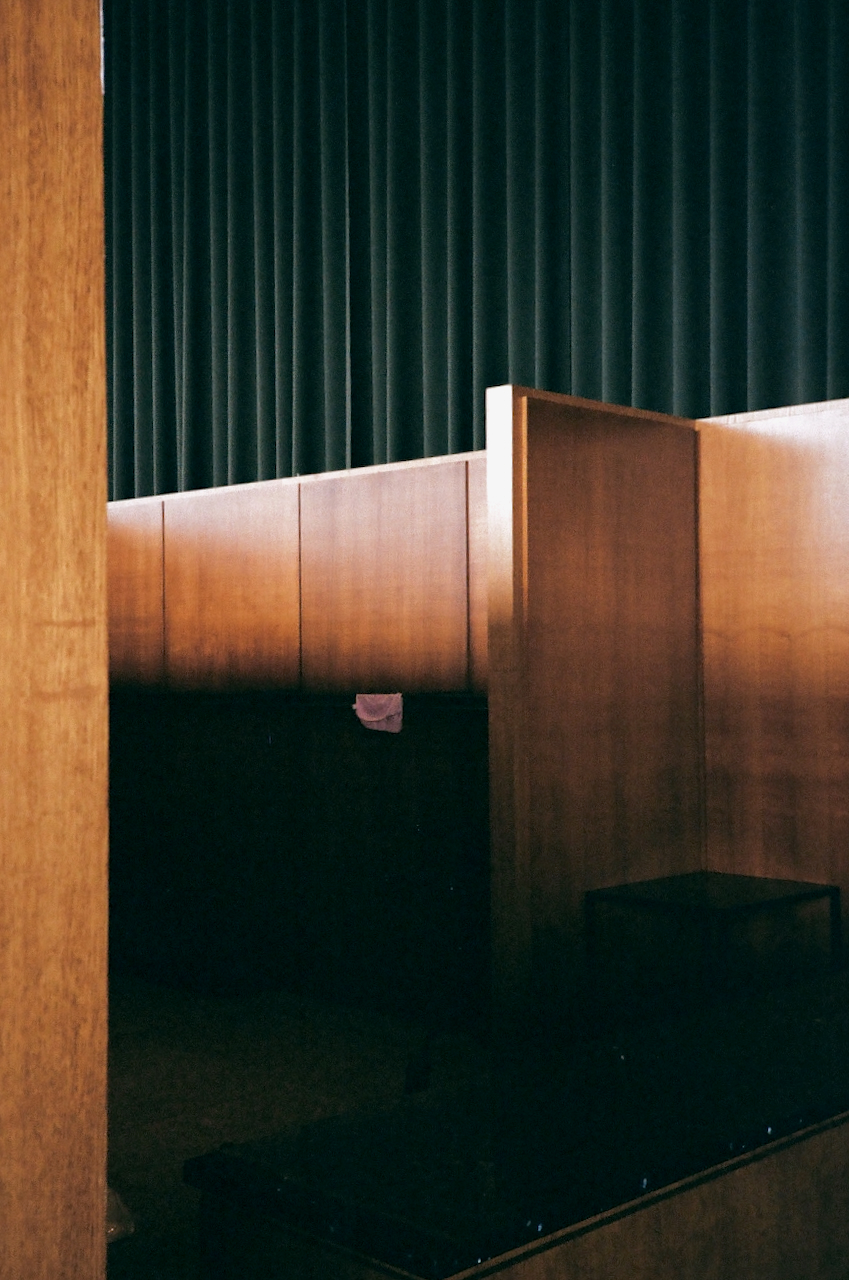 Graph of an interior space with a dark, textured ceiling and a row of wooden benches. The benches are arranged in a long, straight line, and the space appears to be a waiting area or a small auditorium. The lighting is subdued, and the overall atmosphere is quiet and still
