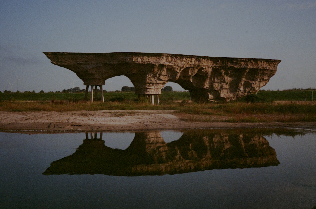 A large, old, stone bridge with multiple arches, reflecting in a calm body of water, in a rural setting