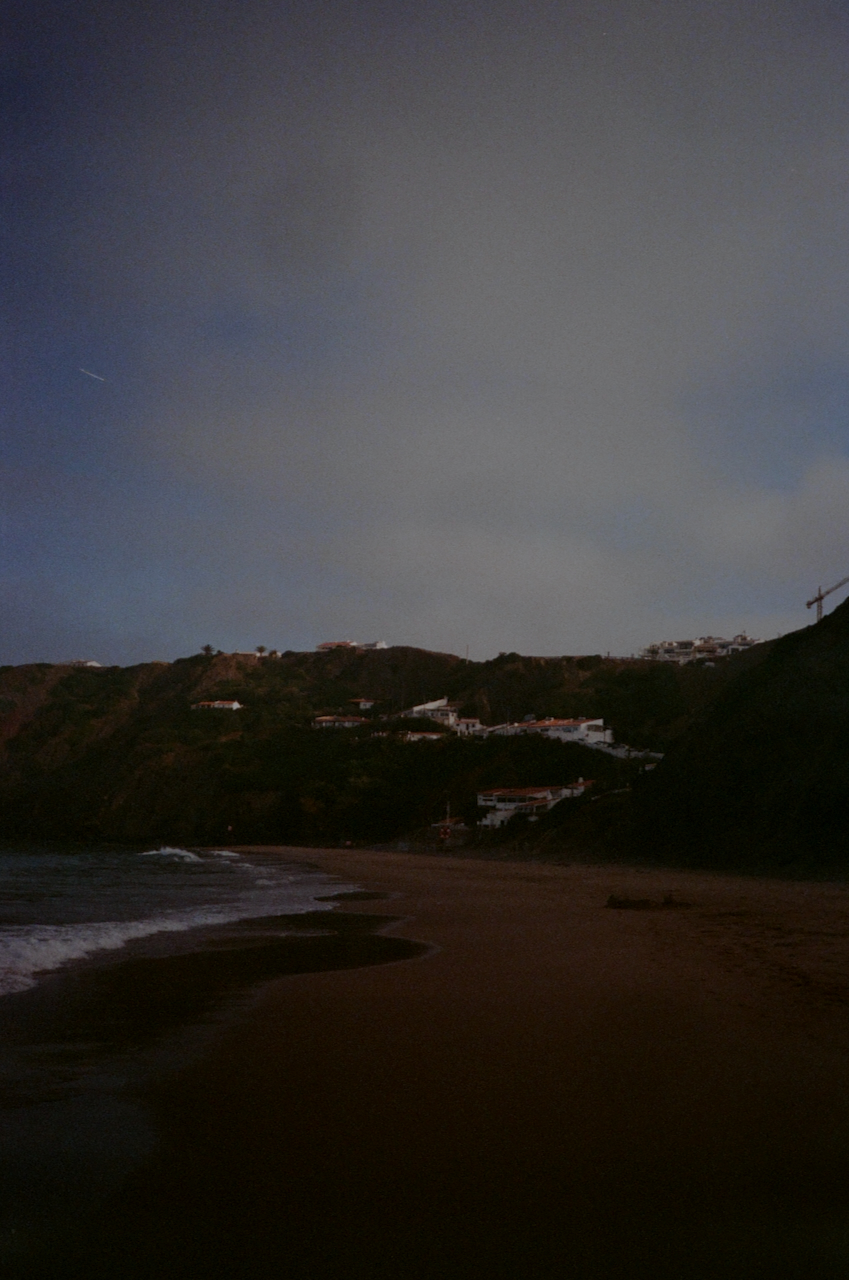 A nighttime beach scene with a dark sky, a house in the distance, and a calm ocean