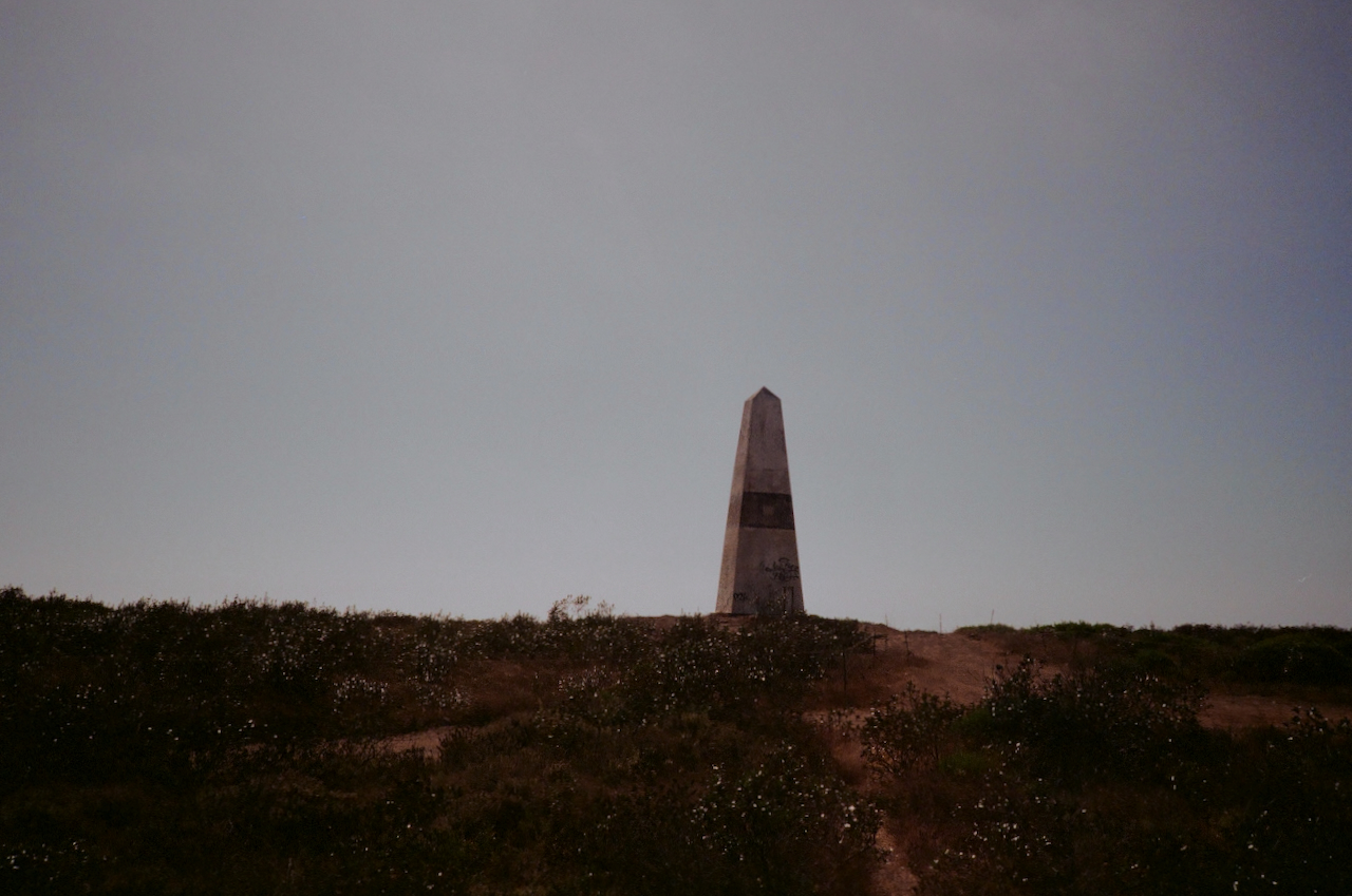 A tall, white, rectangular stone monument stands alone in a field with a clear sky in the background
