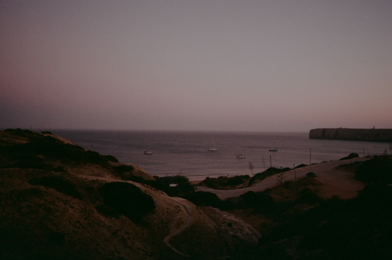 A tranquil beach scene at sunset with a calm ocean, boats, and a coastal town in the background