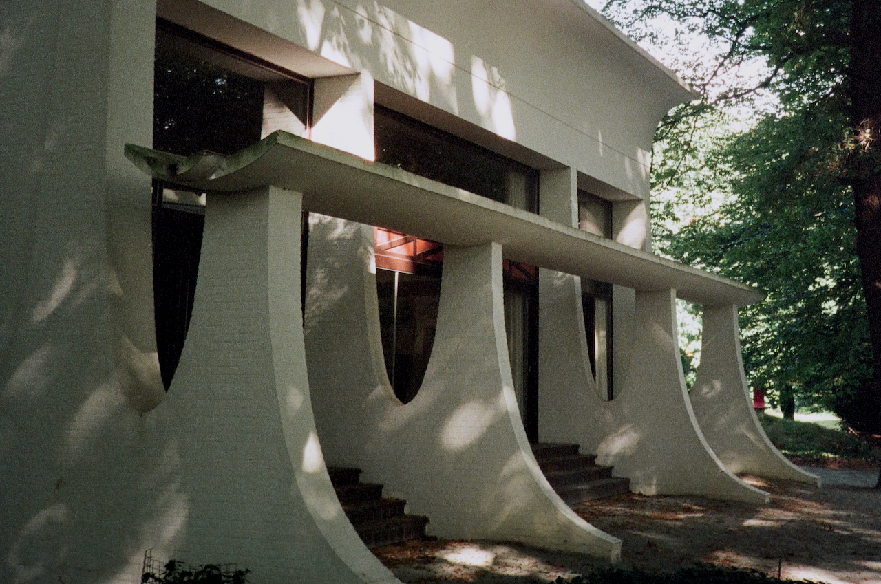 A modern building with a curved roof, set in a park-like environment with trees and a clear sky