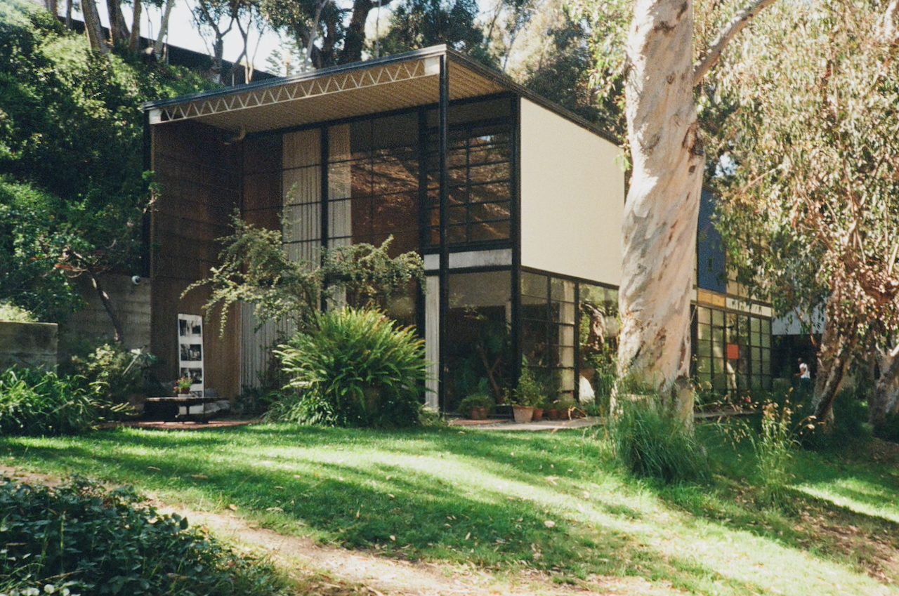 A modern house with a large glass wall, surrounded by a well-maintained garden and trees, on a sunny day